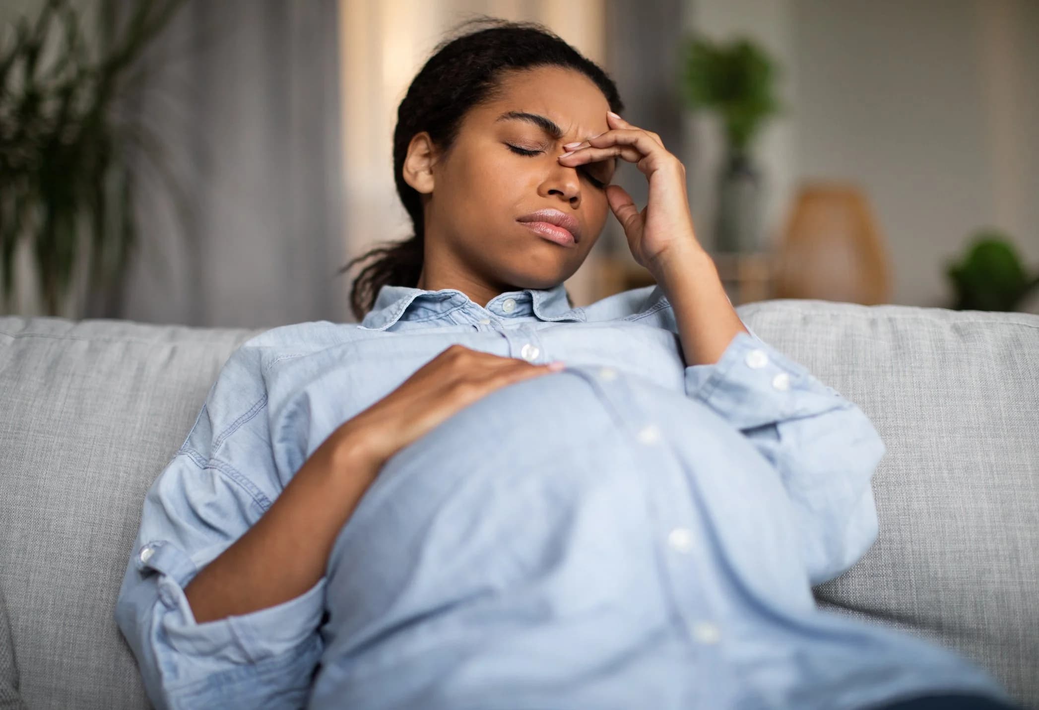 A pregnant woman sitting at home on a sofa with her hand on her forehead, looking tired and showing potential symptoms of high blood pressure.